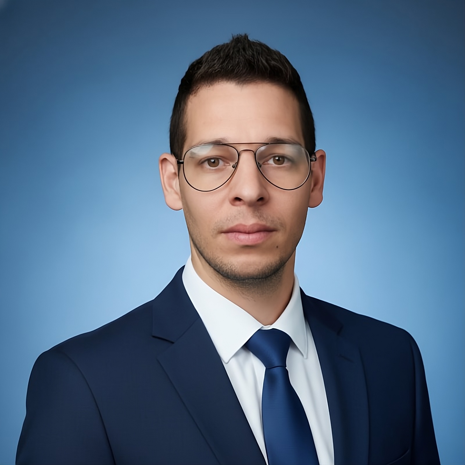 Hombre con gafas y traje azul marino posando frente a un fondo azul claro, fotografía profesional para perfil corporativo.