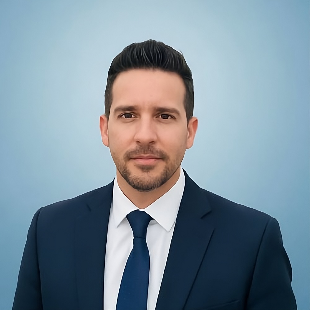 Hombre con traje azul marino y corbata azul, posando frente a un fondo azul claro para fotografía profesional corporativa.